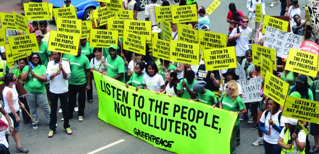 Greenpeace protesters in the street, most have placard and some hold a large banner. The placards and banner all read: 'Listen to the people, not polluters'