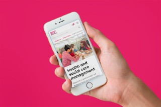 A hand holding an iPhone on a pink background. The iPhone has the Sheffield Hallam University website on it
