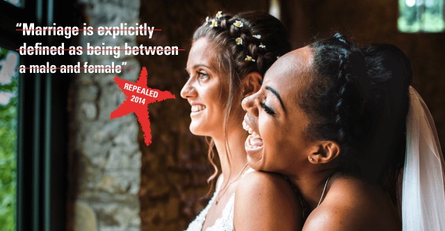 Two brides positioned on the right of an image, both looking left and smiling. One is leaning on the others shoulder. Some crossed out text reads 'Marriage is explicity defined as being between a male and female'. Underneath the text a red star highlights the words 'repealed 2014'.