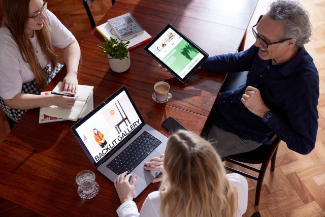 Three people sitting around a wooden table, looking at a tablet and laptop with Backlit Gallery website designs
