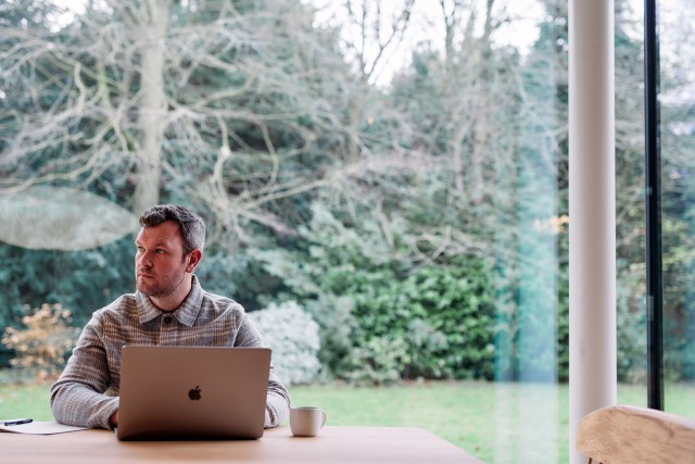 A man sat at a laptop with a coffee. A large glass window and a garden behind