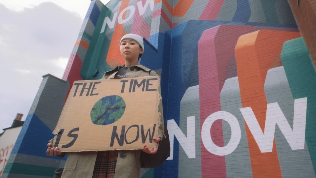 A girl holding a cardboard placard stating 'The Time is Now'. There is a painted picture of the earth in the centre of the placard