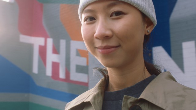 A closeup of a smiling young woman, wearing a wooly hat. A colourful is visible in the background