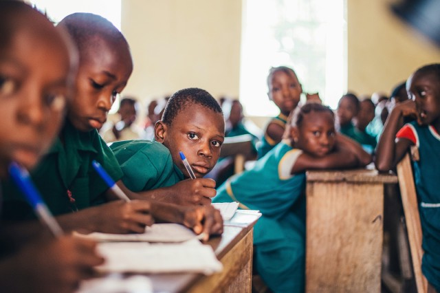 Children at desks in a school, working with blue pens. One child is looking directly at the camera