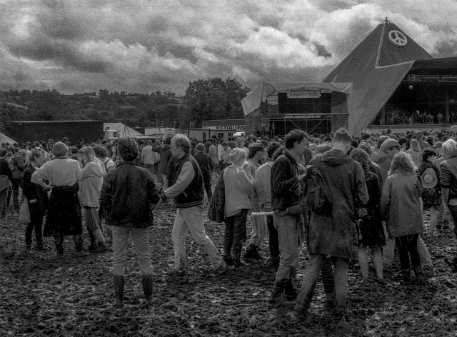 A black and white photograph of a Glastonbury Festival, showing festival goers walking through mud