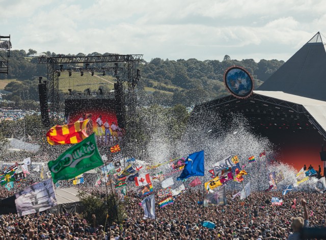A photograph of the Glastonbury Festival Pyramid Stage in the day time taken from a high position, flags and and ticker tape are visible above a large crowd