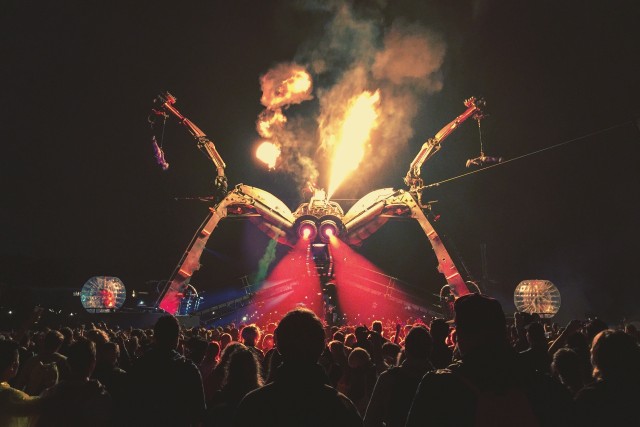 A photograph from Glastonbury Festival - a large robot with two spider-like legs stands over a crowd in the dark. Fire is shooting from it's 'head' and it's 'eyes' light up the crowd with red beams